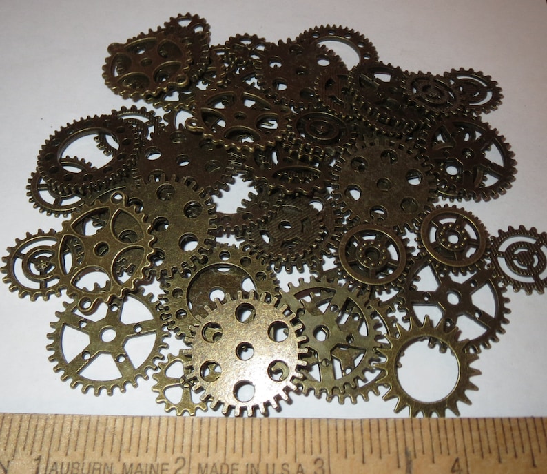 May include: A collection of antique gold-toned metal gears of various sizes. The gears are arranged in a random pattern on a white surface.