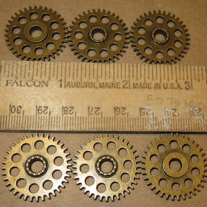 May include: Six gold-colored metal gears with a circular hole in the center and a row of small holes around the outer edge. The gears are arranged in two rows of three, with a ruler underneath.