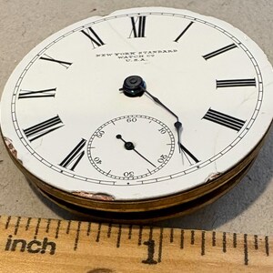 May include: A white pocket watch face with black Roman numeral hour markers and black hands. The watch face has the text "NEW YORK STANDARD WATCH CO. U.S.A." printed on it. The watch face is about 1 inch in diameter.