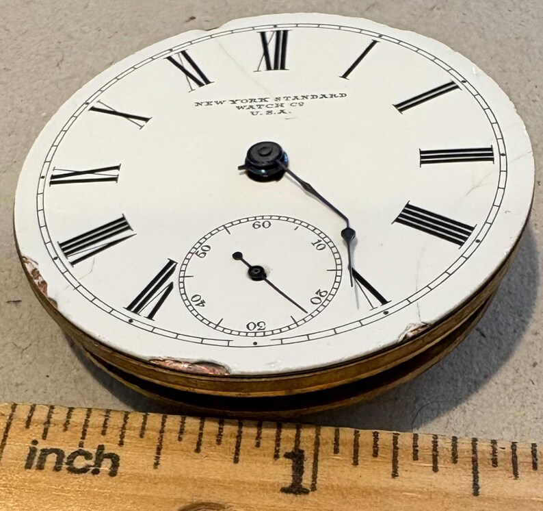 May include: A white pocket watch face with black Roman numeral hour markers and black hands. The watch face has the text "NEW YORK STANDARD WATCH CO. U.S.A." printed on it. The watch face is about 1 inch in diameter.