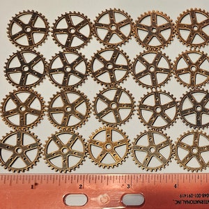 May include: A collection of 18 antique gold-toned metal gears, each with a different design. The gears are arranged in a grid pattern on a white surface. A ruler is included for scale.