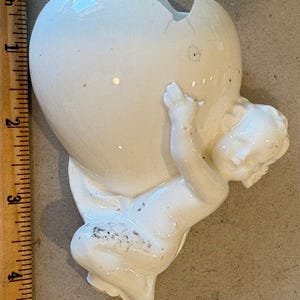 May include: A white ceramic figurine of a cherub holding a large, cracked heart-shaped object. The cherub is detailed with delicate features. The object is approximately 4 inches tall.