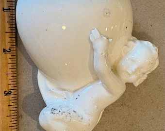 May include: A white ceramic figurine of a cherub holding a large, cracked heart-shaped object. The cherub is detailed with delicate features. The object is approximately 4 inches tall.