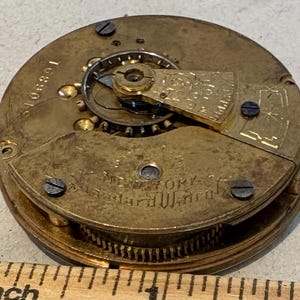 May include: Close-up of a gold-toned pocket watch movement with the inscription "New York City Alexander Watch Co." visible. The watch has a number "106891" on the side.