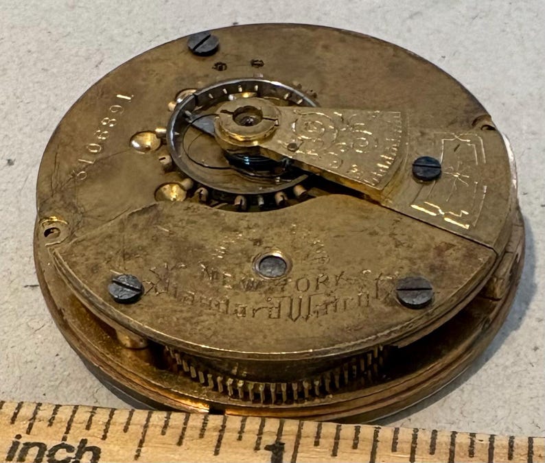 May include: Close-up of a gold-toned pocket watch movement with the inscription "New York City Alexander Watch Co." visible. The watch has a number "106891" on the side.