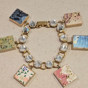 May include: A gold chain bracelet with clear crystal beads and square charms. The charms are made of paper with different patterns and colors, including green, blue, pink, and white.