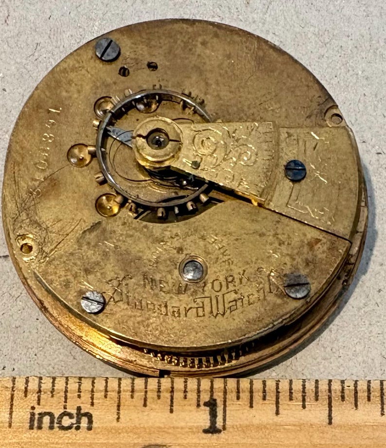 May include: Close-up of a gold-toned pocket watch movement with the inscription "New York Standard Watch Co." visible. The watch movement is disassembled and shows the gears and other internal components.