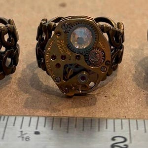 May include: Three rings featuring circular watch movement faces with exposed gears and metallic components, set on ornate bronze-toned filigree bands. A metal ruler is visible at the bottom, showing the rings measure approximately 1 inch in diameter.