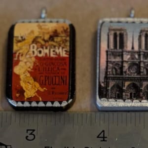 May include: Six small rectangular and oval pendants featuring vintage-style art prints, including a black cat, Notre Dame, and Moulin Rouge, arranged above a metal ruler showing the pendants are approximately 1 inch in height and width.
