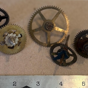 May include: Five metallic clock gears of varying sizes and designs arranged above a metal ruler. The gears feature brass and dark steel components with central shafts and teeth. The ruler shows measurements in inches, with the gears spanning from 1 to 5 inches.