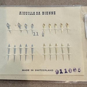 May include: A collection of twenty watch hands arranged on a cream-colored card inside a clear plastic sleeve. The card features the text AIGUILLA SA BIENNE, the number 11 1/2, and MADE IN SWITZERLAND. The watch hands are silver and gold-toned.