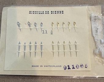 May include: A collection of twenty watch hands arranged on a cream-colored card inside a clear plastic sleeve. The card features the text AIGUILLA SA BIENNE, the number 11 1/2, and MADE IN SWITZERLAND. The watch hands are silver and gold-toned.