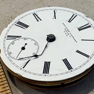 May include: A white pocket watch face with black Roman numeral hour markers and black minute markers. The watch face has the text "NEW YORK STANDARD WATCH CO. U.S.A." printed on it.