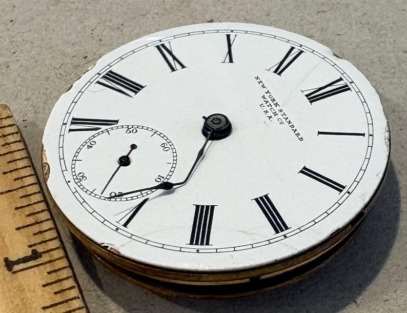 May include: A white pocket watch face with black Roman numeral hour markers and black minute markers. The watch face has the text "NEW YORK STANDARD WATCH CO. U.S.A." printed on it.