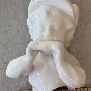 May include: A small, off-white ceramic figurine of a cherub, with arms raised to the face. The figurine is approximately 1 inch tall. The cherub has a serene expression and is wearing a head covering. The surface is glossy.
