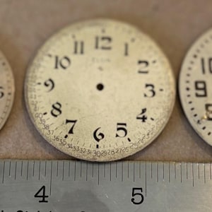May include: Three circular white watch dials with black Arabic numerals arranged along a metal ruler. The left dial has a sub-dial, the center dial features a date track around the edge, and the right dial includes a small sub-dial at the three o'clock position.