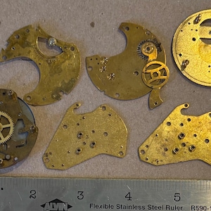 May include: A collection of five gold-toned brass watch movement plates and bridges with various gears and holes, arranged above a metal ruler showing measurements in inches, with the largest circular plate measuring approximately 1.5 inches.