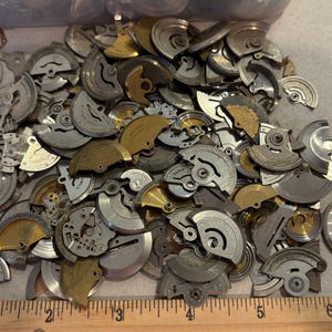 May include: A large pile of assorted silver and gold watch parts. The parts are various shapes and sizes, and some have intricate designs.