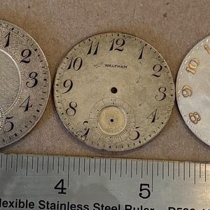 May include: Three circular silver-toned Waltham watch dials with black or gold numerals and sub-dials, positioned above a stainless steel ruler showing measurements in inches, with the dials ranging from approximately 1.25 to 1.5 inches in diameter.
