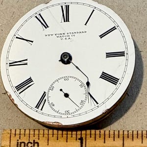 May include: Close-up of a white pocket watch face with black Roman numeral hour markers and black hands. The watch face reads "NEW YORK STANDARD WATCH CO. U.S.A.".