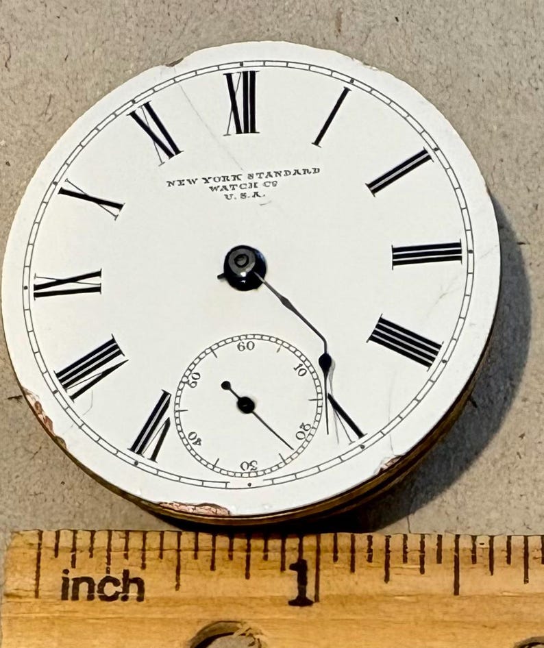 May include: Close-up of a white pocket watch face with black Roman numeral hour markers and black hands. The watch face reads "NEW YORK STANDARD WATCH CO. U.S.A.".