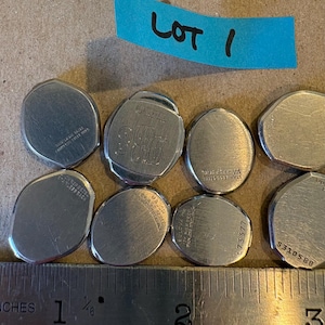 May include: A collection of eight silver-toned metal watch case backs of various shapes and sizes, some featuring engraved text and serial numbers, arranged above a metal ruler showing measurements in inches for scale, with a blue label reading LOT 1.