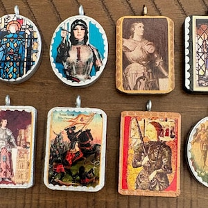 May include: Eight decorative pendants featuring historical and religious imagery, including depictions of Joan of Arc in armor, stained glass patterns, and battle scenes. Each pendant has a metal bail and a scalloped or smooth frame edge.