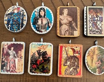 May include: Eight decorative pendants featuring historical and religious imagery, including depictions of Joan of Arc in armor, stained glass patterns, and battle scenes. Each pendant has a metal bail and a scalloped or smooth frame edge.