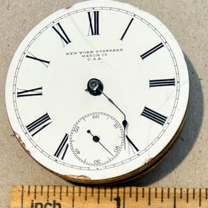 May include: White pocket watch face with black Roman numeral hour markers and black minute markers. The watch face reads "NEW YORK STANDARD WATCH CO. U.S.A."