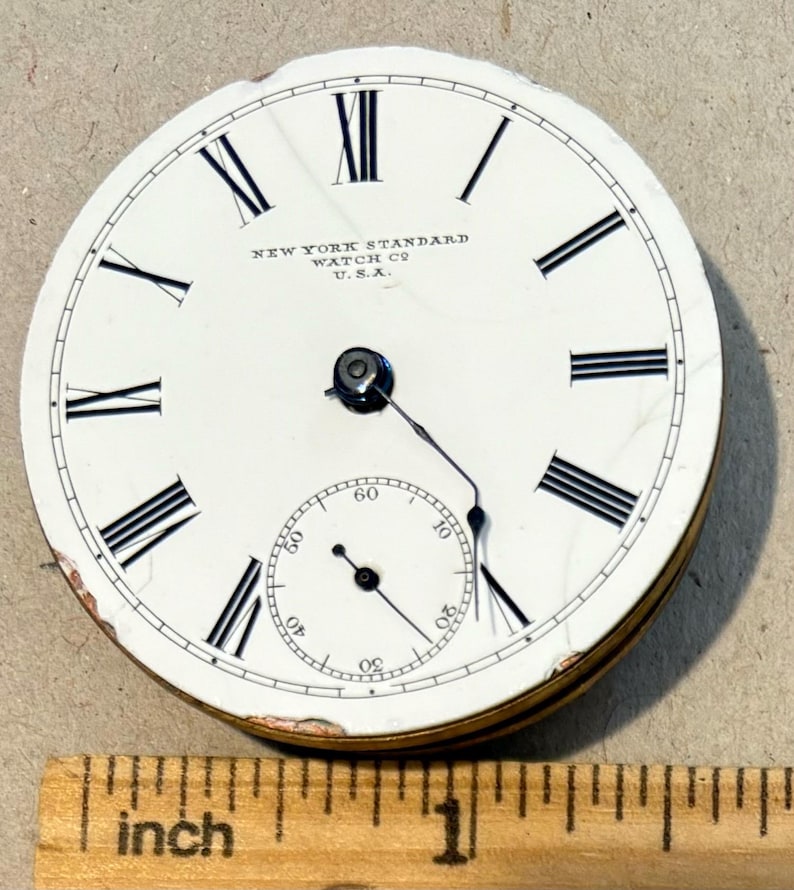 May include: White pocket watch face with black Roman numeral hour markers and black minute markers. The watch face reads "NEW YORK STANDARD WATCH CO. U.S.A."