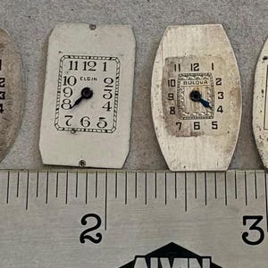 May include: Six vintage watch faces of various shapes and designs are displayed. The faces are off-white with black numerals and hands. A ruler with inch measurements is at the bottom. The faces measure approximately 1 inch to 1.5 inches in length.