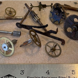 May include: A collection of assorted metal clock and watch parts, including various gears, pinions, and levers, arranged above a stainless steel ruler that measures up to 6 inches, showing the scale of these small mechanical components.