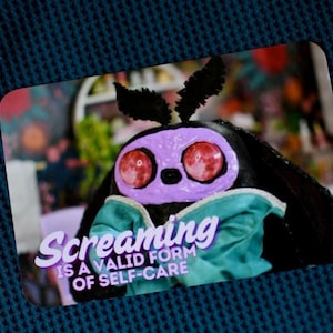 May include: A purple and black creature with large red eyes and a teal bow tie. The creature is on a card with the text "Screaming is a valid form of self-care".