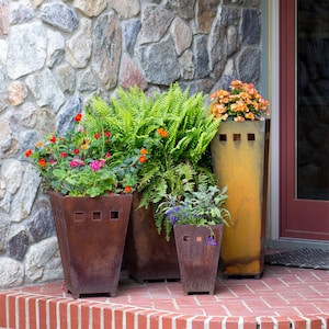 May include: A collection of outdoor planters filled with vibrant flowers and greenery. The planters are made of weathered metal with square cutouts, and are arranged on a brick surface in front of a stone wall.