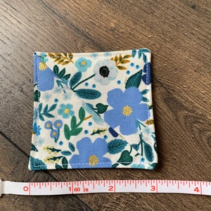May include: A square coaster with a floral pattern in shades of blue, green, and yellow on a white background. The coaster measures 1.6 inches in diameter.