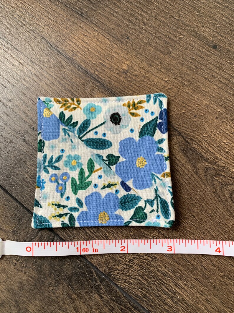 May include: A square coaster with a floral pattern in shades of blue, green, and yellow on a white background. The coaster measures 1.6 inches in diameter.
