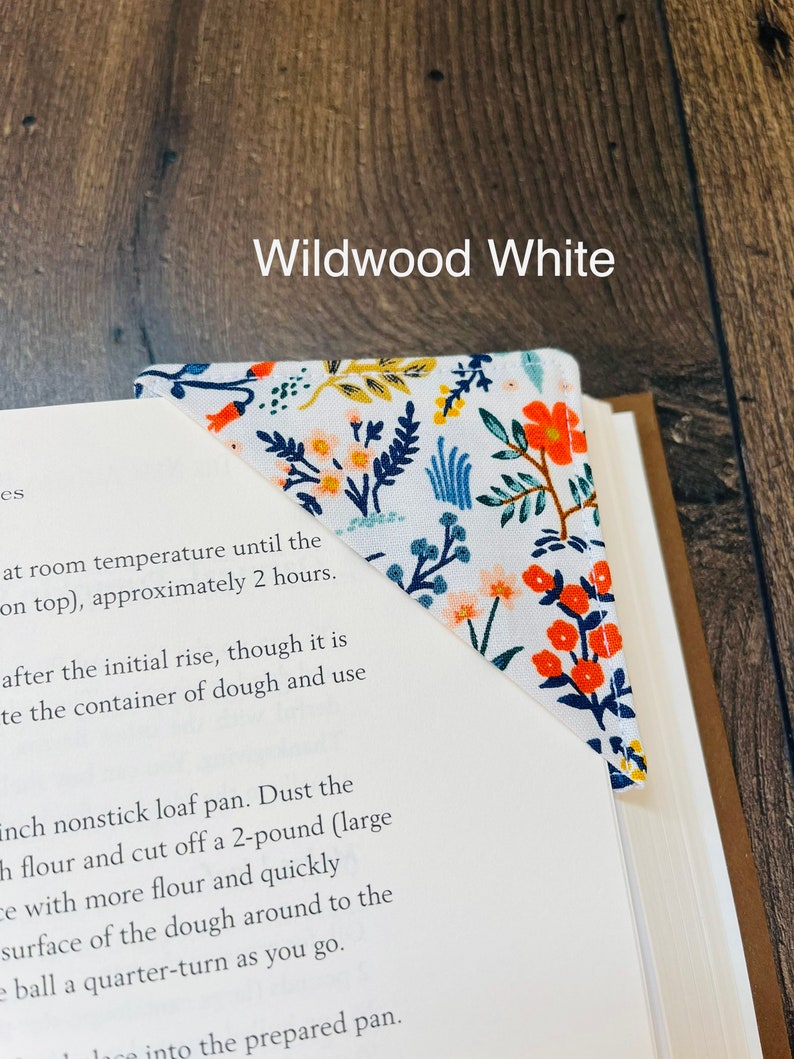 May include: A white fabric bookmark with a floral pattern in shades of blue, orange, and yellow. The bookmark is folded into a triangle shape and has the text "Wildwood White" printed on the top.