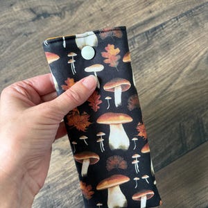 May include: A black fabric glasses case featuring a repeating pattern of mushrooms and autumn leaves in shades of brown, orange, and white. The case is secured with a light green button.