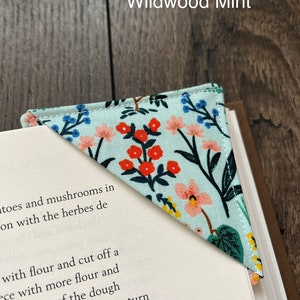 May include: A light blue bookmark with a floral pattern in shades of pink, red, yellow, and green. The bookmark is shaped like a triangle and has a white background. The text "Wildwood Mint" is printed in black at the top of the image.