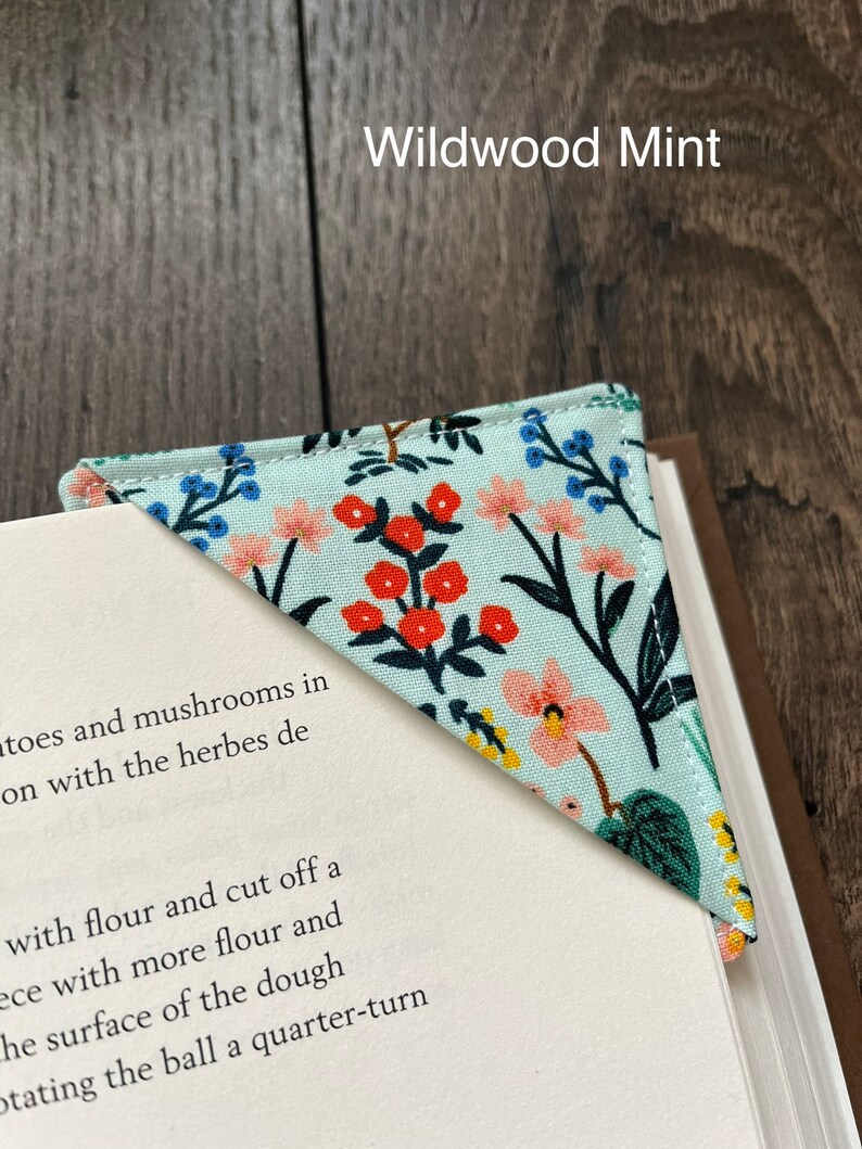 May include: A light blue bookmark with a floral pattern in shades of pink, red, yellow, and green. The bookmark is shaped like a triangle and has a white background. The text "Wildwood Mint" is printed in black at the top of the image.