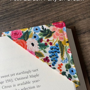 May include: A floral fabric bookmark with a white background and a variety of colorful flowers, including pink, red, blue, and yellow. The bookmark is in the shape of a triangle and has a white stitched edge. The text "Petit Garden Party on Cream" is visible at the top of the image.