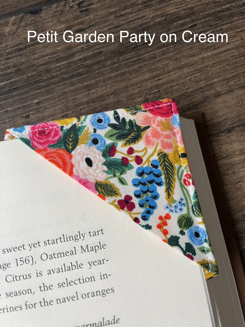 May include: A floral fabric bookmark with a white background and a variety of colorful flowers, including pink, red, blue, and yellow. The bookmark is in the shape of a triangle and has a white stitched edge. The text "Petit Garden Party on Cream" is visible at the top of the image.