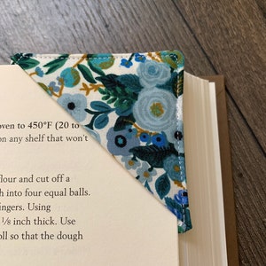 May include: A blue and white fabric bookmark with a floral pattern. The bookmark is folded into a triangle shape and is placed on a book. The text "Petit Garden Party Blue" is visible at the top of the image.