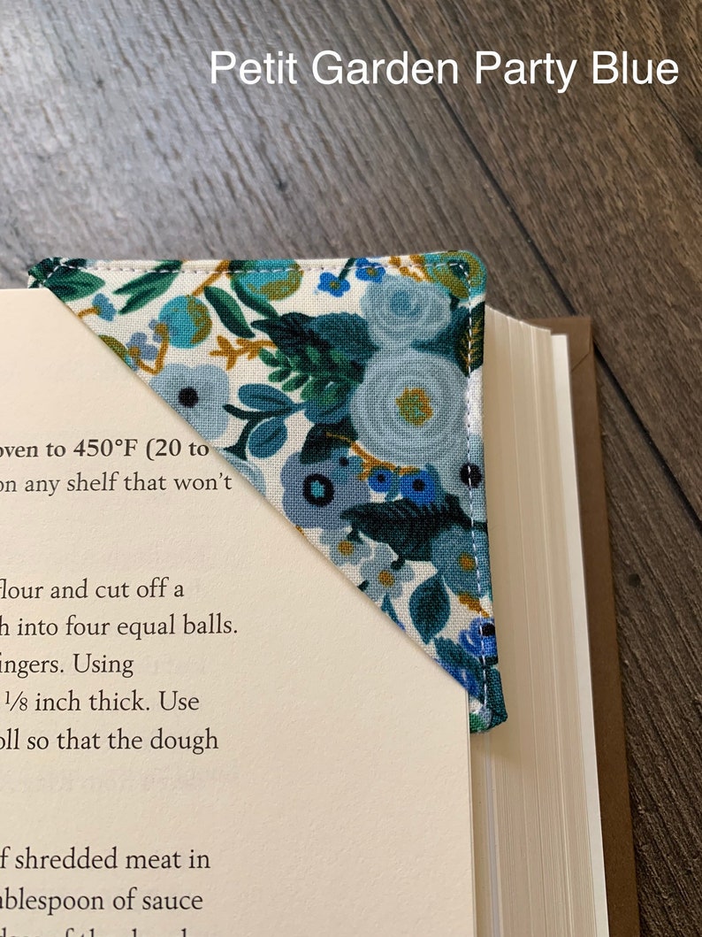 May include: A blue and white fabric bookmark with a floral pattern. The bookmark is folded into a triangle shape and is placed on a book. The text "Petit Garden Party Blue" is visible at the top of the image.