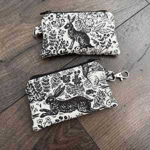 May include: Two small, rectangular zippered pouches with a black and white rabbit and floral print. One pouch has a small loop, the other has a metal clasp. The pouches are on a dark wood surface.