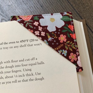 May include: A burgundy fabric bookmark with a floral pattern featuring white and pink flowers. The bookmark is shaped like a triangle and has a white border. The text "Wild Rose Burgundy" is printed at the top of the image.