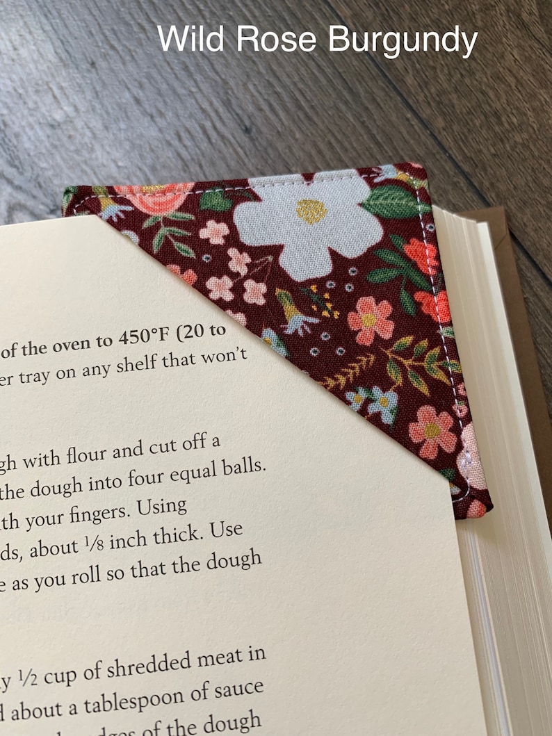 May include: A burgundy fabric bookmark with a floral pattern featuring white and pink flowers. The bookmark is shaped like a triangle and has a white border. The text "Wild Rose Burgundy" is printed at the top of the image.