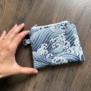 May include: A small fabric zipper pouch featuring a blue and white Japanese-style wave pattern. The rectangular pouch includes a white zipper closure along the top edge and a small metal D-ring attached to the side for hanging or attachment purposes.