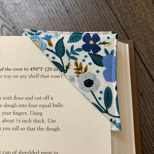 May include: A white fabric bookmark with a blue and green floral pattern. The bookmark is folded into a triangle shape and has the text "Wild Rose Blue" printed on it.