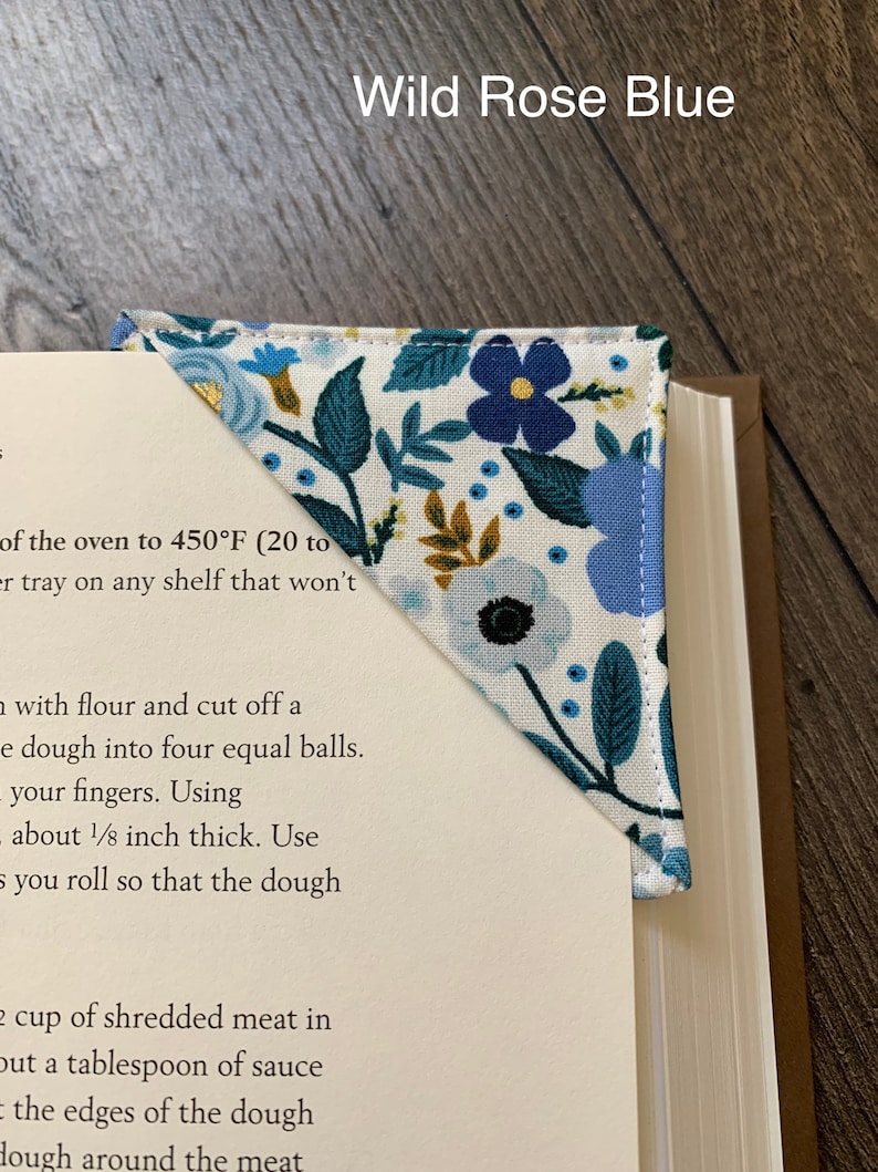 May include: A white fabric bookmark with a blue and green floral pattern. The bookmark is folded into a triangle shape and has the text "Wild Rose Blue" printed on it.